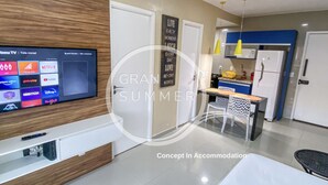 A modern hotel room with a flat-screen TV, a wooden accent wall, a kitchen area with a refrigerator, and a dining table with chairs. - Gran Summer Concept In Accommodation (Fortaleza)