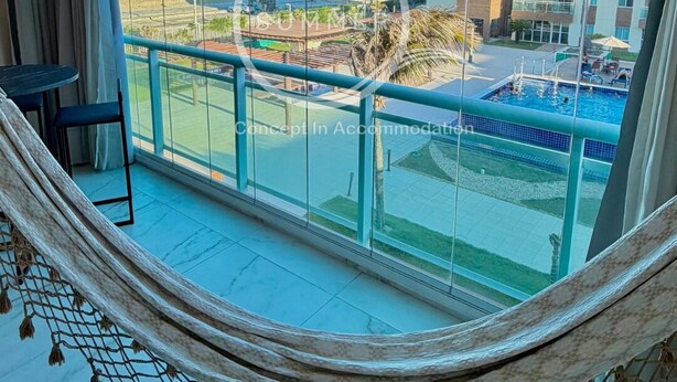 Un balcón con vista a la playa y al área de la piscina, una hamaca y una pequeña mesa.