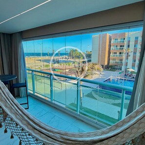 Un balcón con vista a la playa y al área de la piscina, una hamaca y una pequeña mesa.