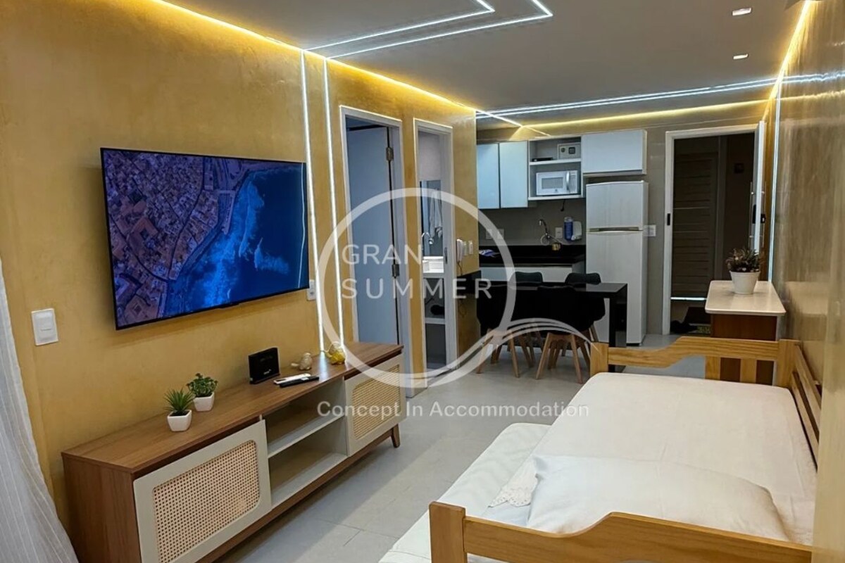 A modern bedroom with a bed, a TV mounted on the wall, a small desk, and a kitchenette in the background.
