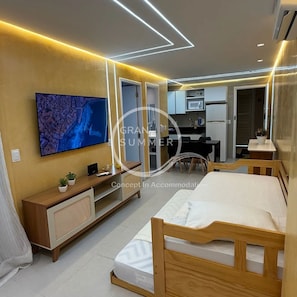 Un dormitorio moderno con una cama, un televisor montado en la pared, un escritorio pequeño y una kitchenette al fondo.