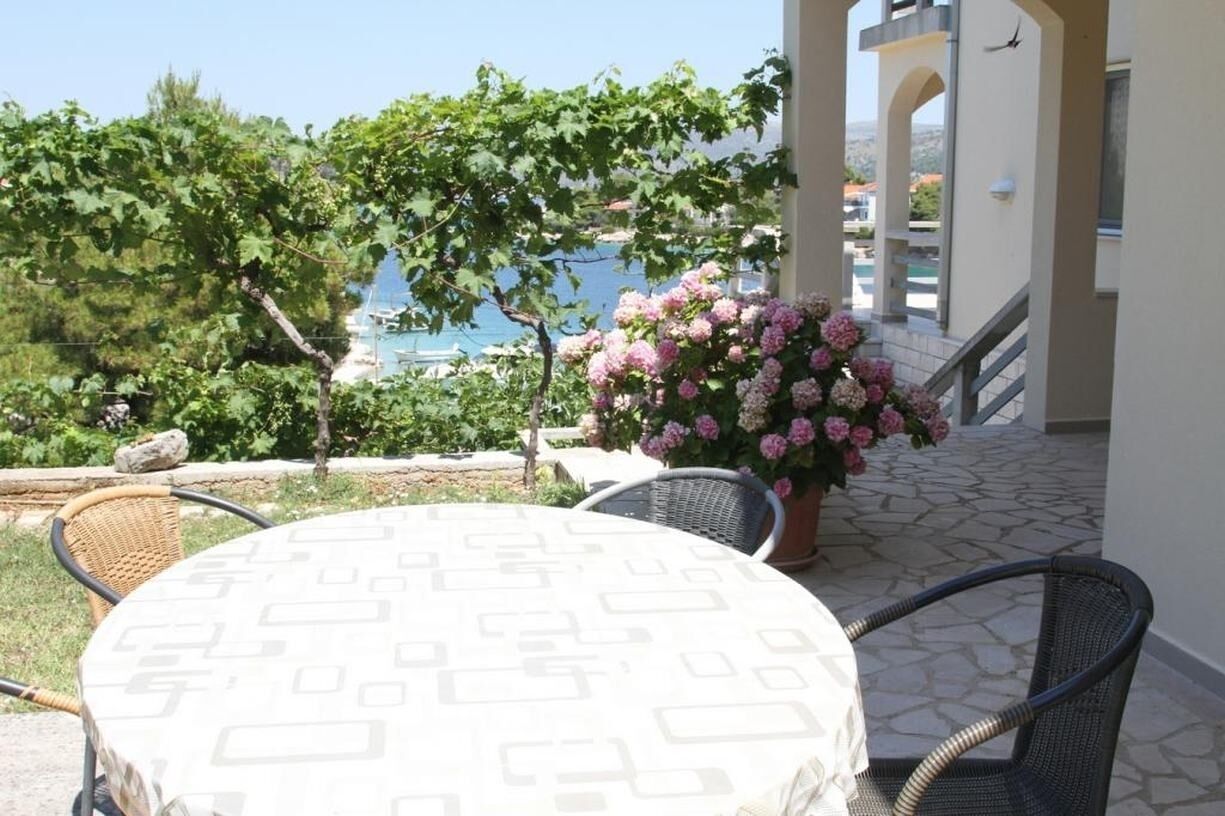 An outdoor patio with a round table and chairs, a potted plant with pink flowers, and a view of the sea and buildings in the background.