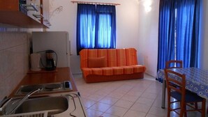 Ein kompakter Wohnbereich mit Küchenzeile, einer Couch, einem Esstisch mit Stühlen und einem Fenster mit blauen Vorhängen.