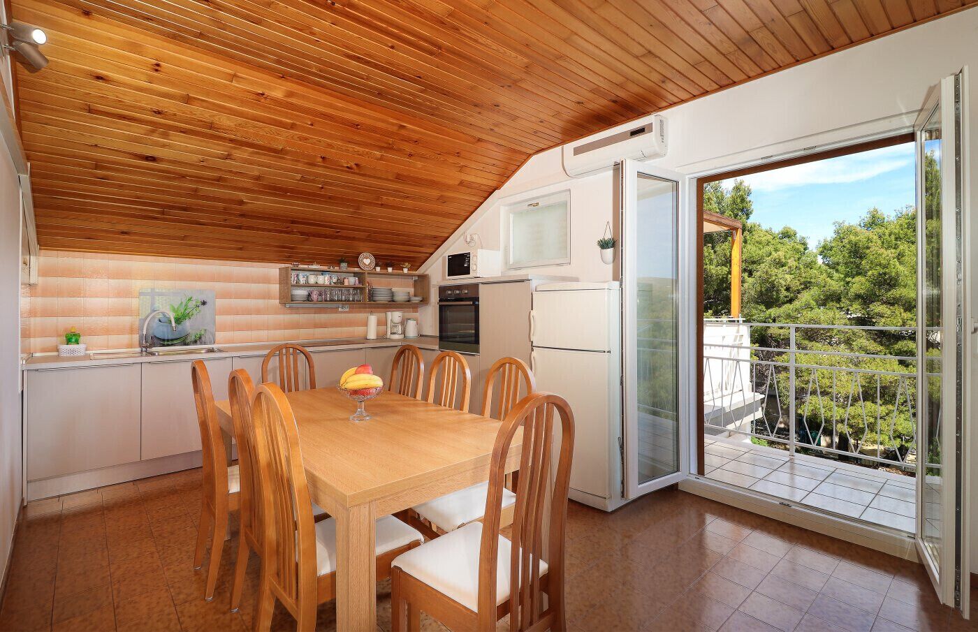 A modern kitchen with wooden ceiling, a dining table with chairs, a refrigerator, and a view of greenery through a glass door.
