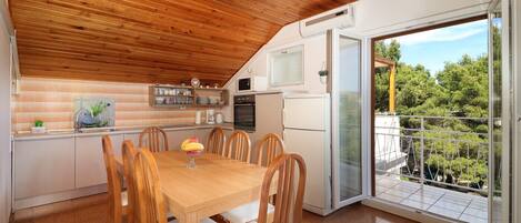 A modern kitchen with wooden ceiling, a dining table with chairs, a refrigerator, and a view of greenery through a glass door.