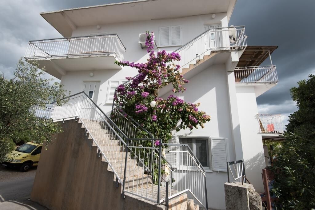 A two-story building with a staircase leading to the upper floor, surrounded by greenery and a parked car.