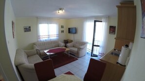 Ein Wohnzimmer mit einer Couch, einem Couchtisch und einem Fernseher. Eine Tür führt zu einem Balkon.