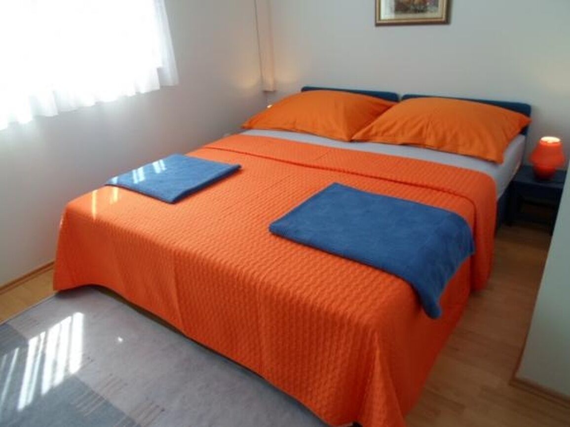A double bed with orange bedding and blue pillows, a bedside table with a lamp, and a window with curtains.