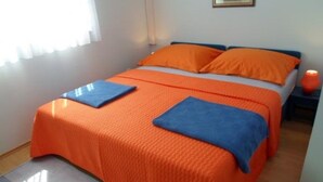 A double bed with orange bedding and blue pillows, a bedside table with a lamp, and a window with curtains.