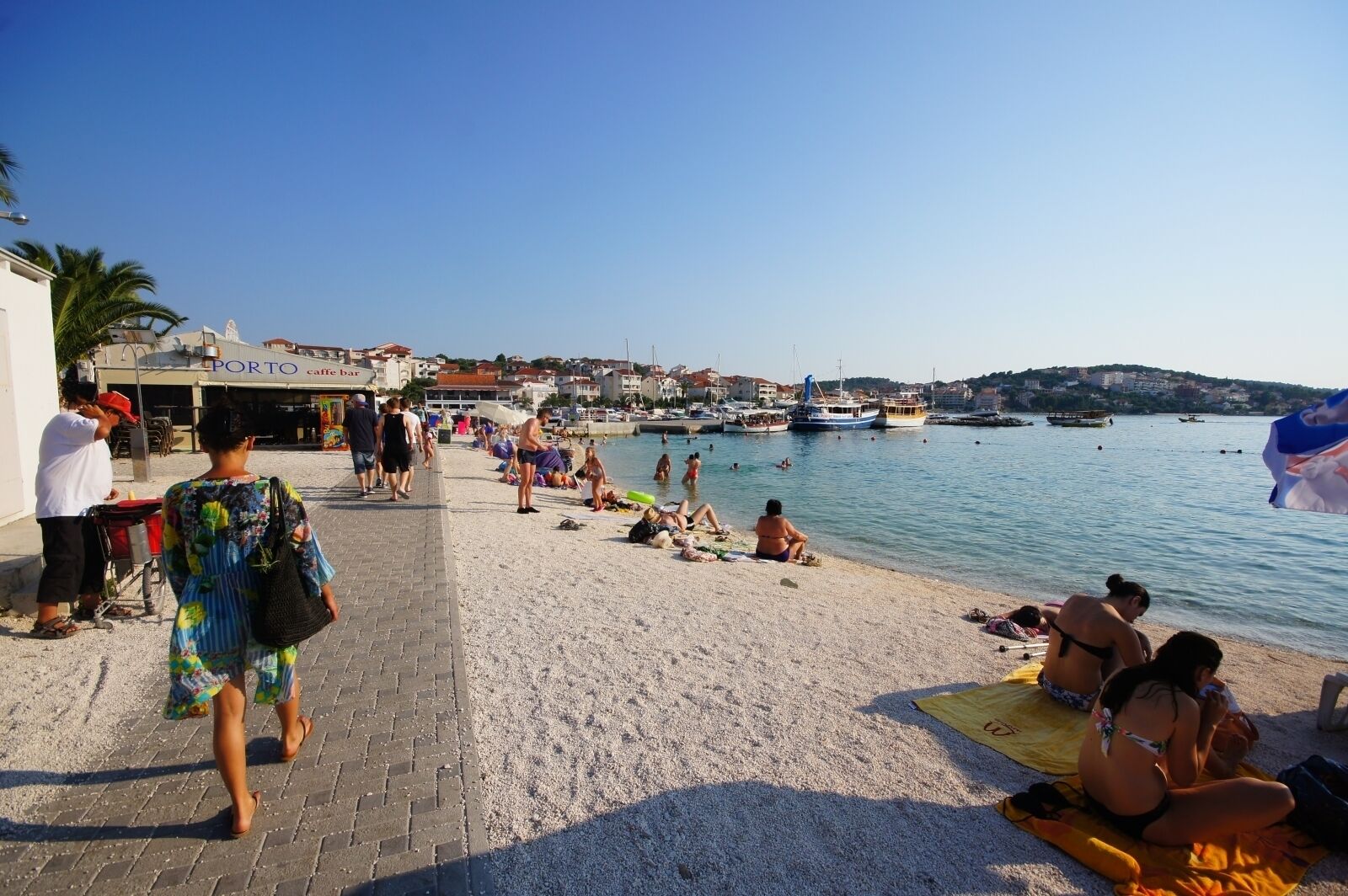 Eine Strandpromenade mit einem Café, Personen, die sonnenbaden, und Booten im Wasser.