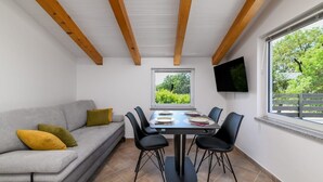Een kamer met een grijze bank, een eettafel met stoelen, een televisie aan de muur en een raam met uitzicht op groen.