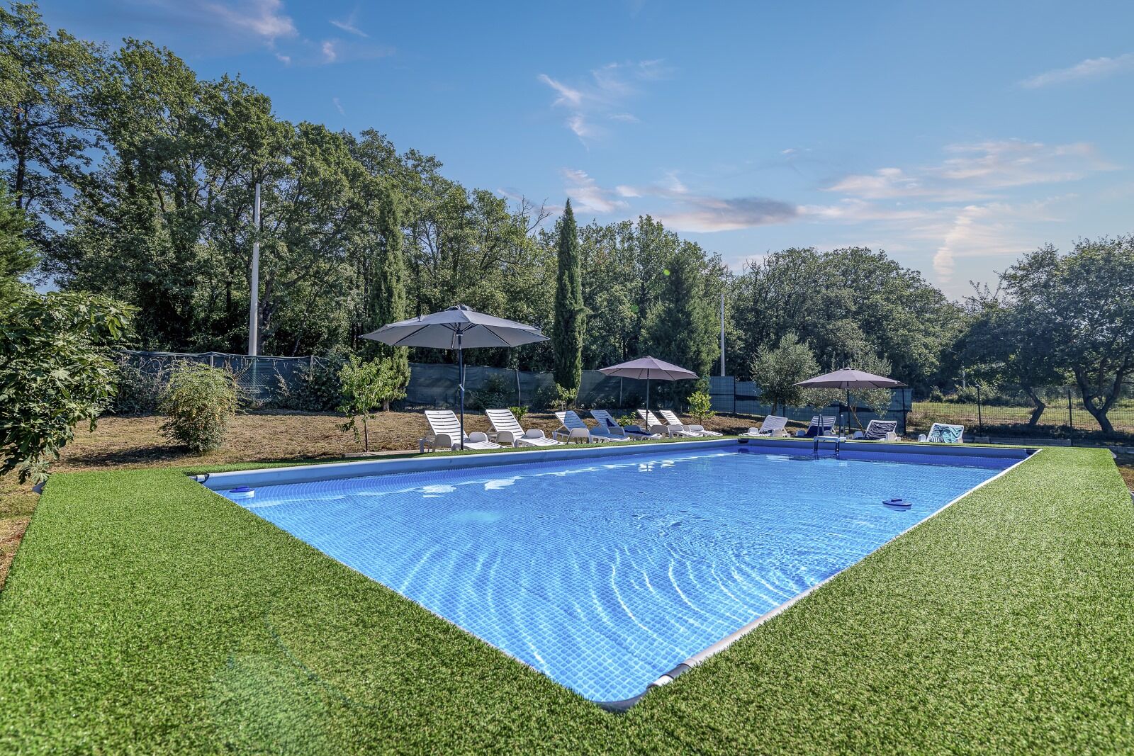 En rektangulær swimmingpool med flisekant, omgivet af en velplejet græsplæne. Der er liggestole og parasoller langs poolen, og en lille bygning med tag i baggrunden.