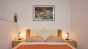 A neatly made bed with a quilted orange bedspread, two pillows, and two bedside lamps on wooden nightstands. A framed picture of a snowy square fountain is hung on the wall above the bed.