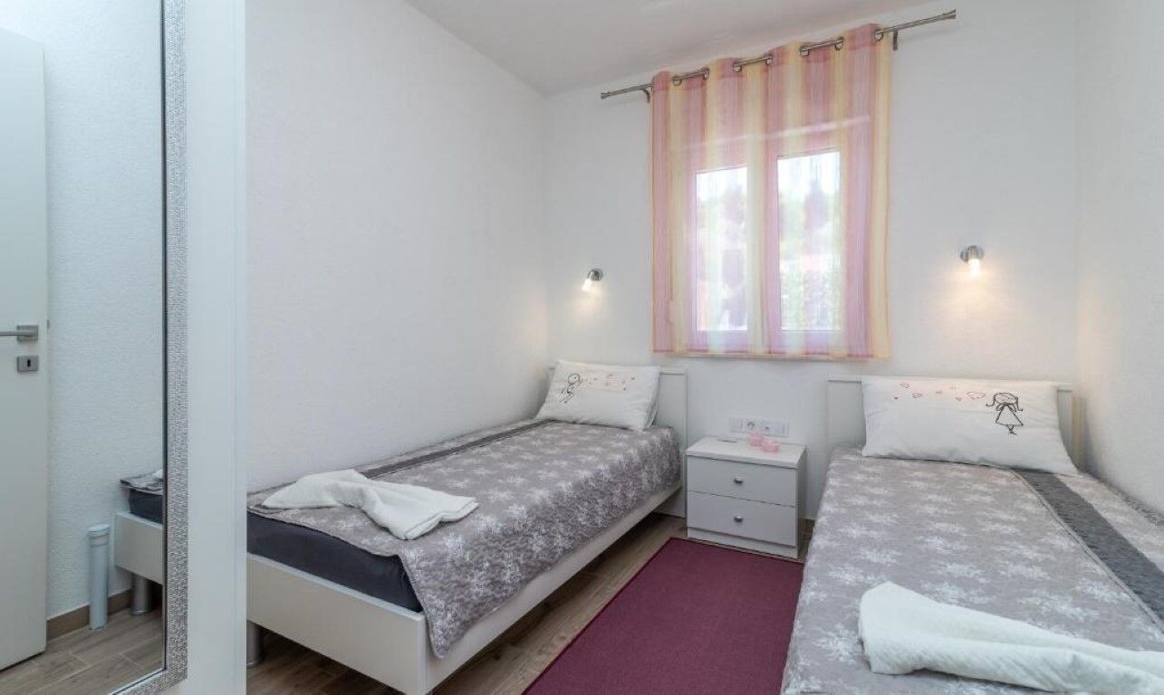 A small, clean room with two single beds, a nightstand, a window with curtains, and a door.