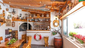 A rustic interior with wooden furniture, shelves with various items, a fireplace, and a view of flowers outside.