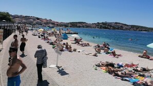 A beach with people sunbathing and swimming, a clear sky, and buildings in the background.