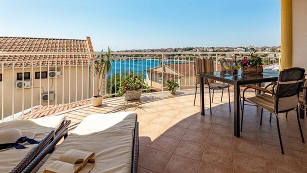 A balcony with a bed, a table, and chairs, overlooking a swimming pool and cityscape.