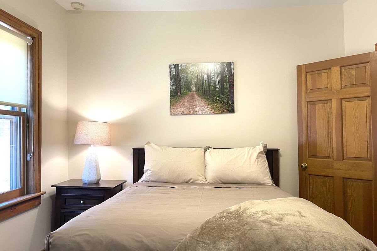 A bedroom with a bed, a nightstand, a lamp, a door, and a wall-mounted picture of a forest path.