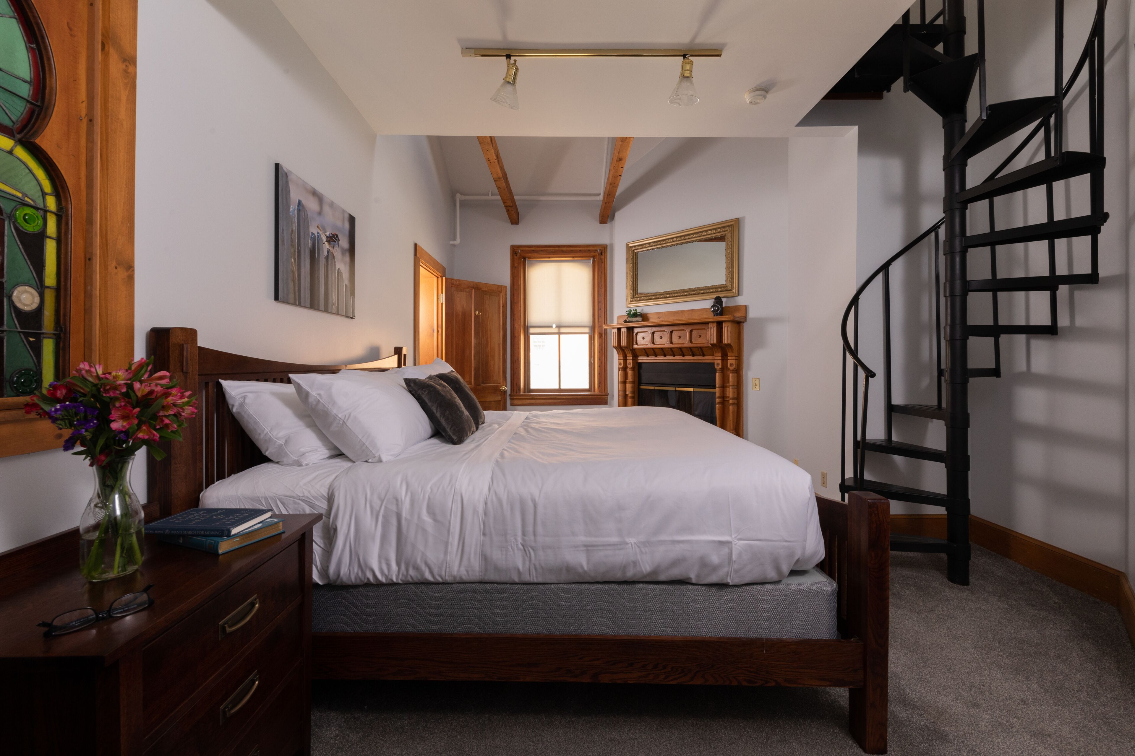 Un dormitorio con una cama de madera, una mesita de noche con un florero, una chimenea y una escalera de caracol.