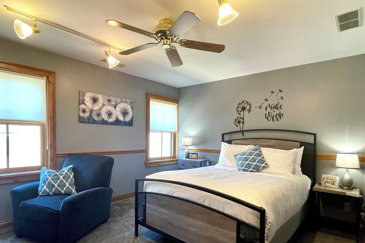 A bedroom with a bed, a chair, a ceiling fan, and a nightstand with a lamp.