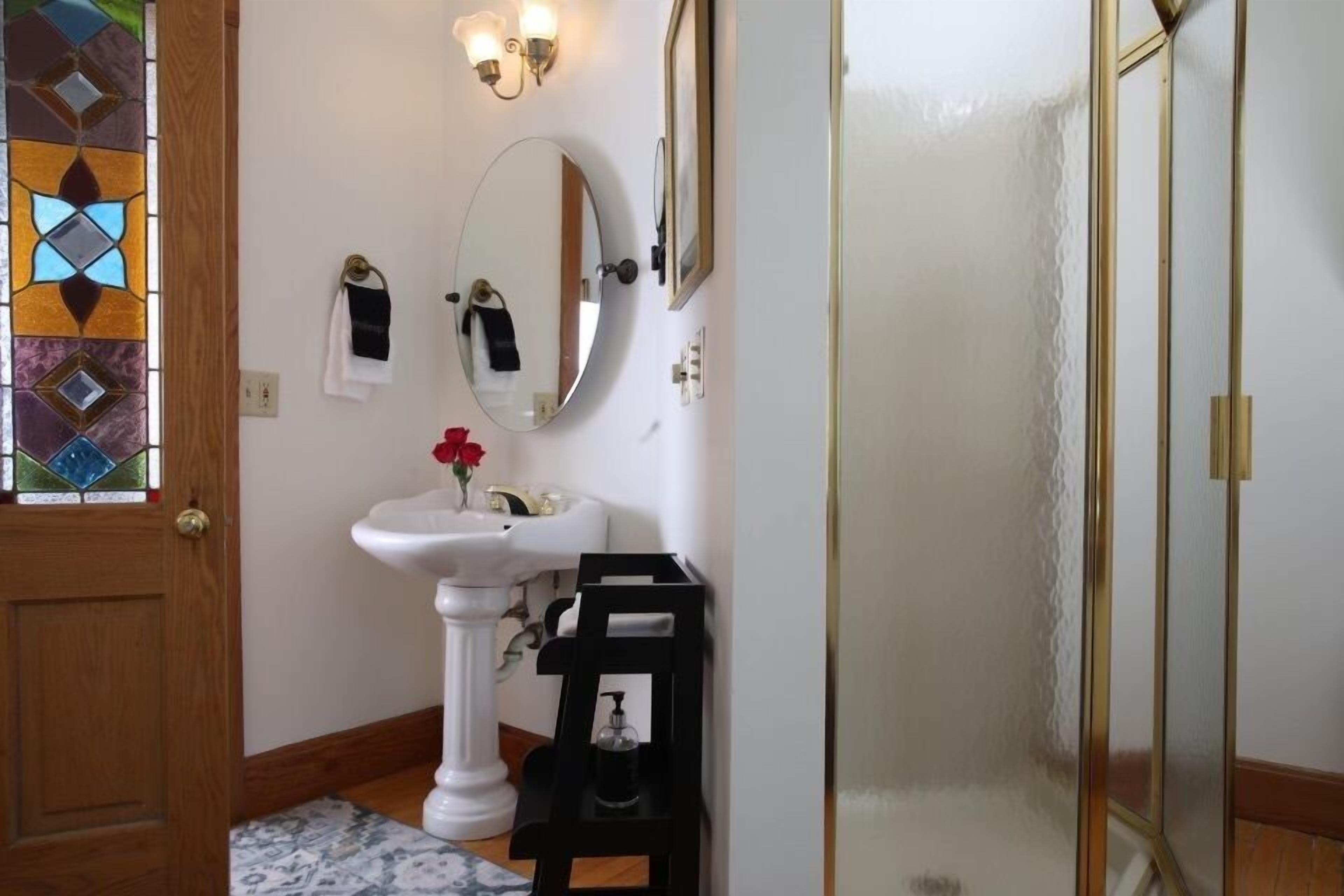 Un baño con un lavamanos pedestal blanco, un espejo, una cabina de ducha con mampara de vidrio y una puerta de madera.