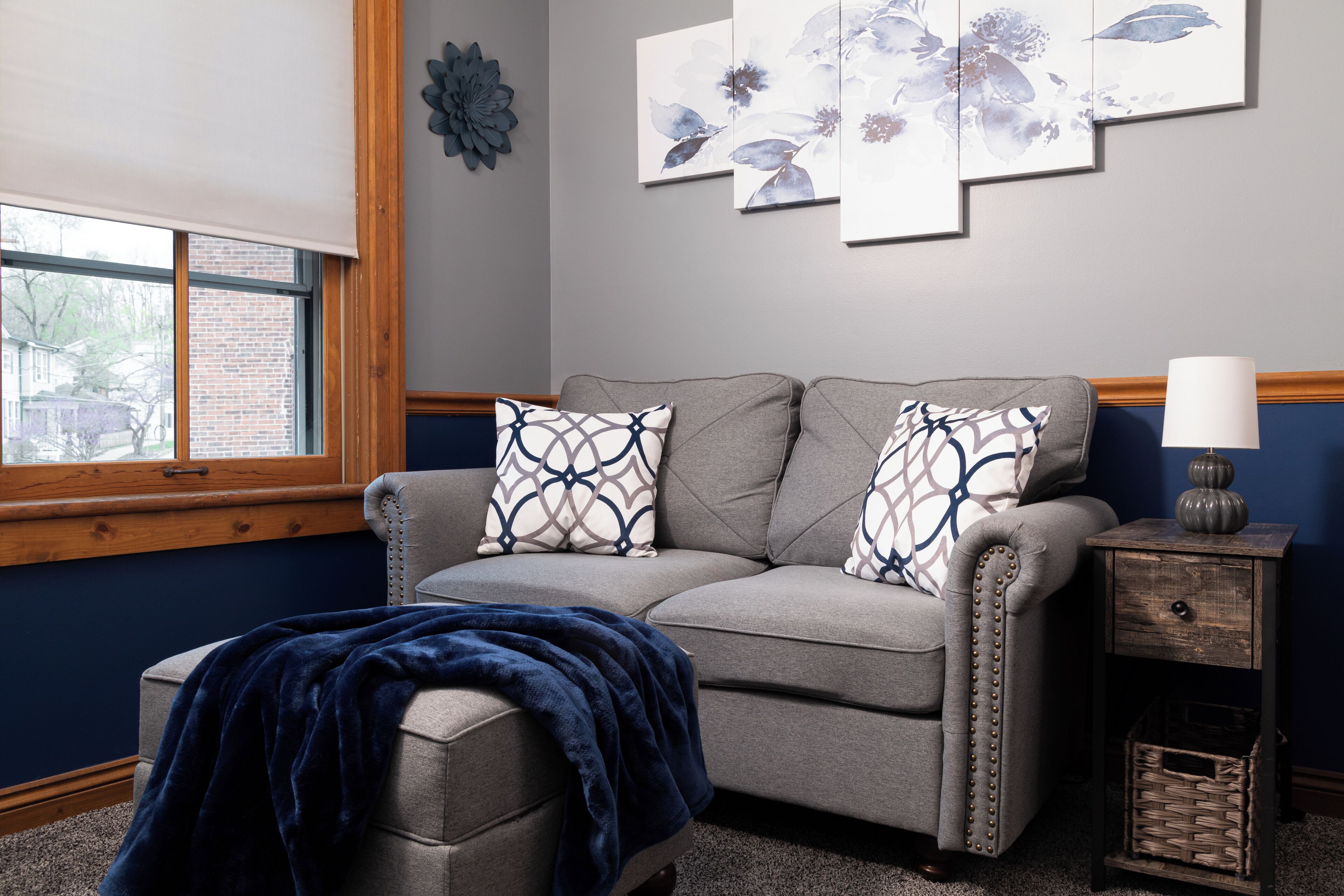 A living room with a grey sofa, patterned cushions, a blue throw blanket, a side table with a lamp, and wall art.