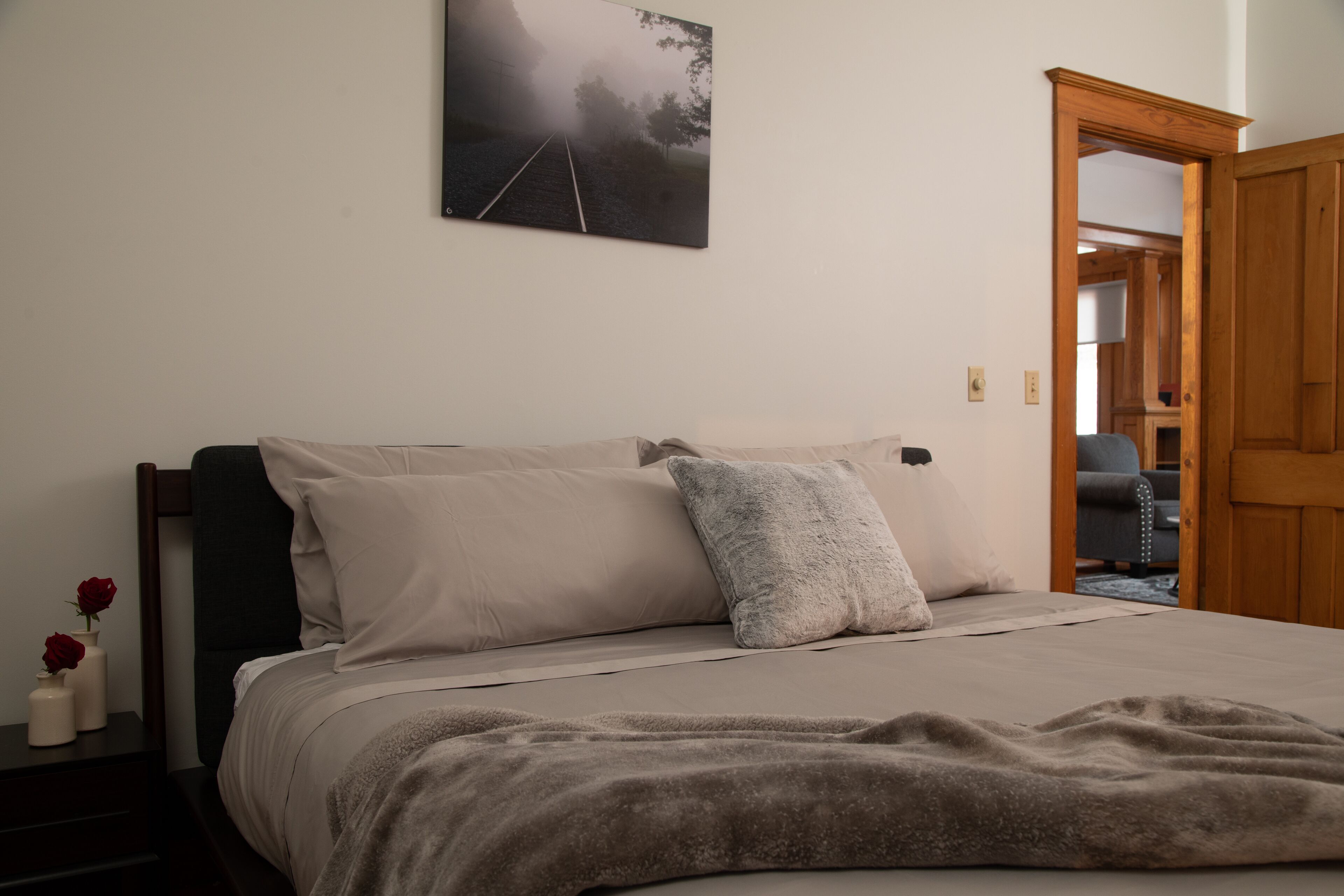 A bedroom with a bed, pillows, a blanket, a nightstand with a vase of flowers, and a painting on the wall.