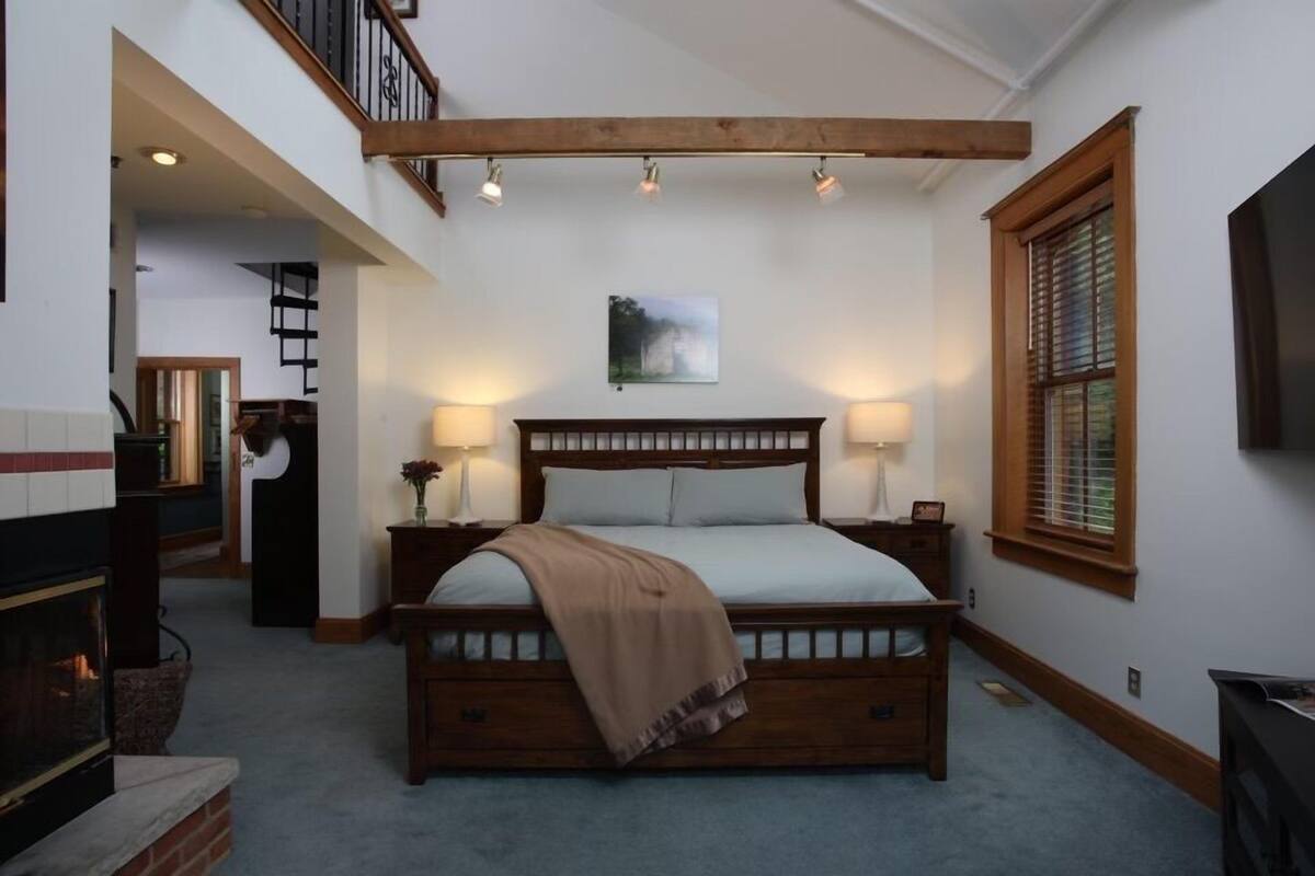 A bedroom with a large bed, two bedside lamps, a fireplace, and a window with blinds.