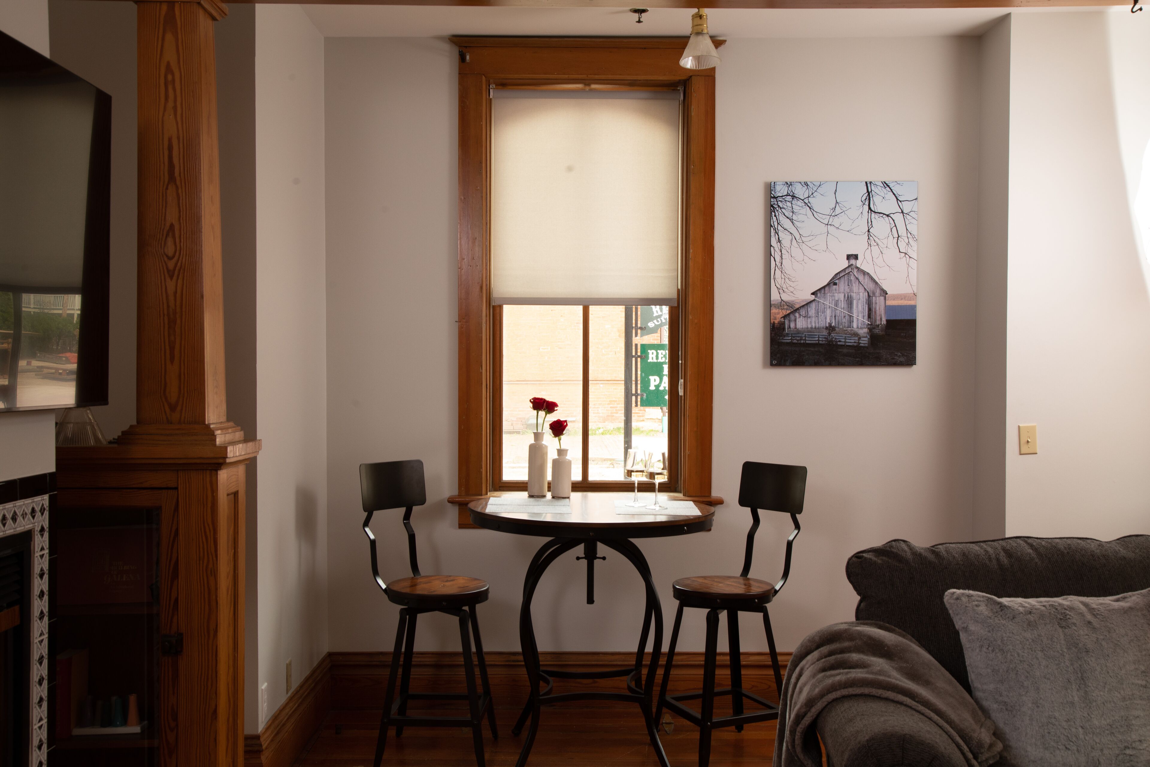 A dining area with a round table, two chairs, a window with a view, and a painting on the wall.