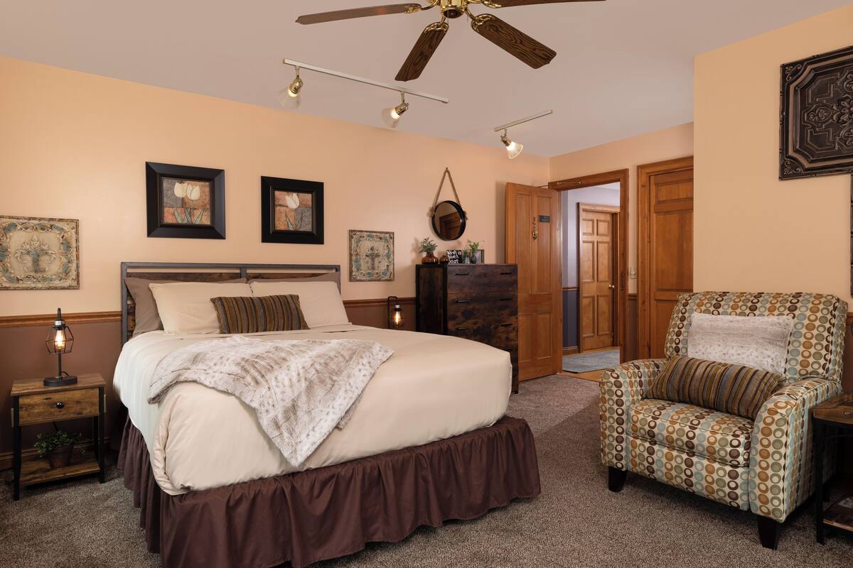 A bedroom with a large bed, a chair, a ceiling fan, and artwork on the walls.