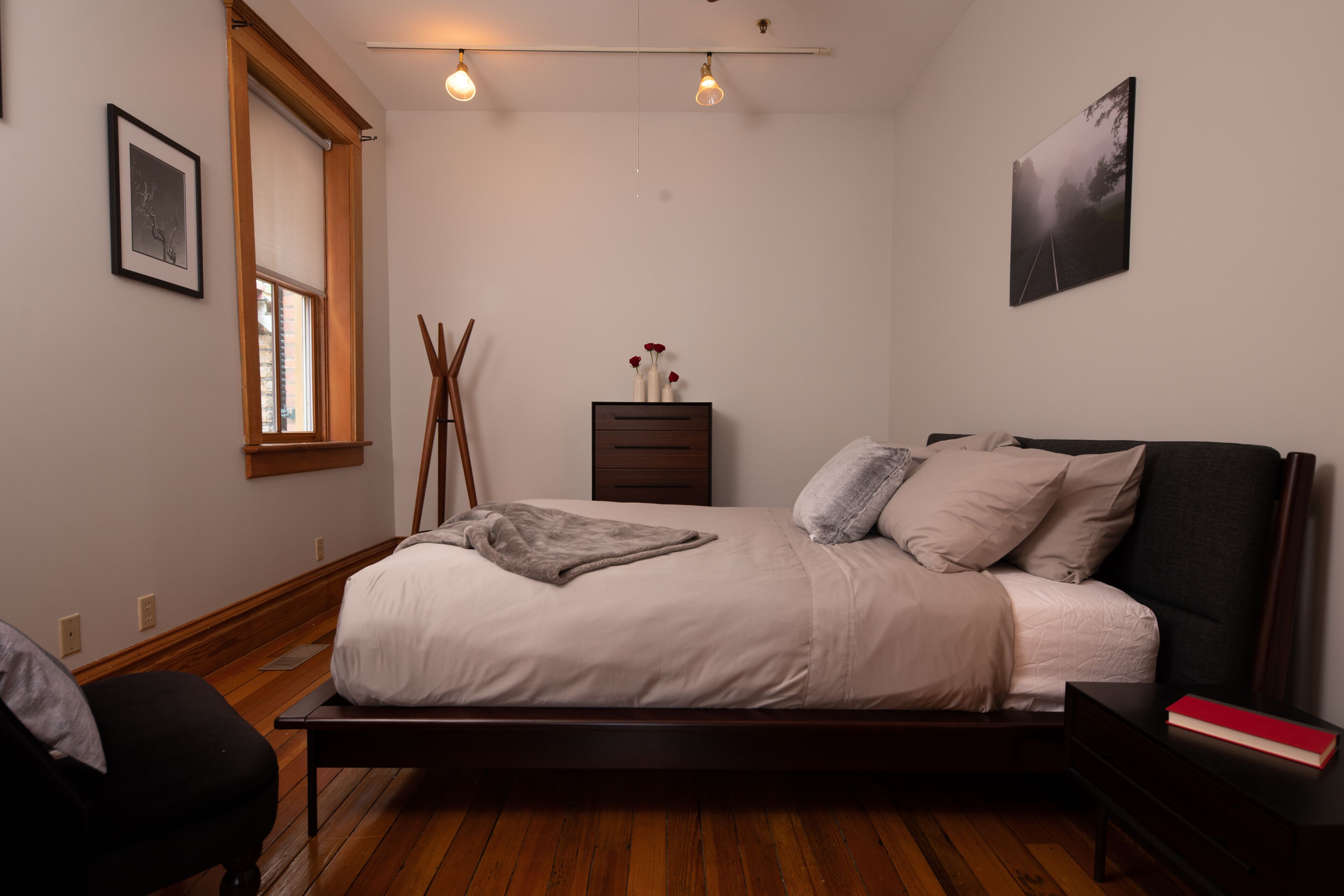 A bedroom with a wooden floor, a bed with white bedding, a nightstand, a dresser, and a window with blinds.