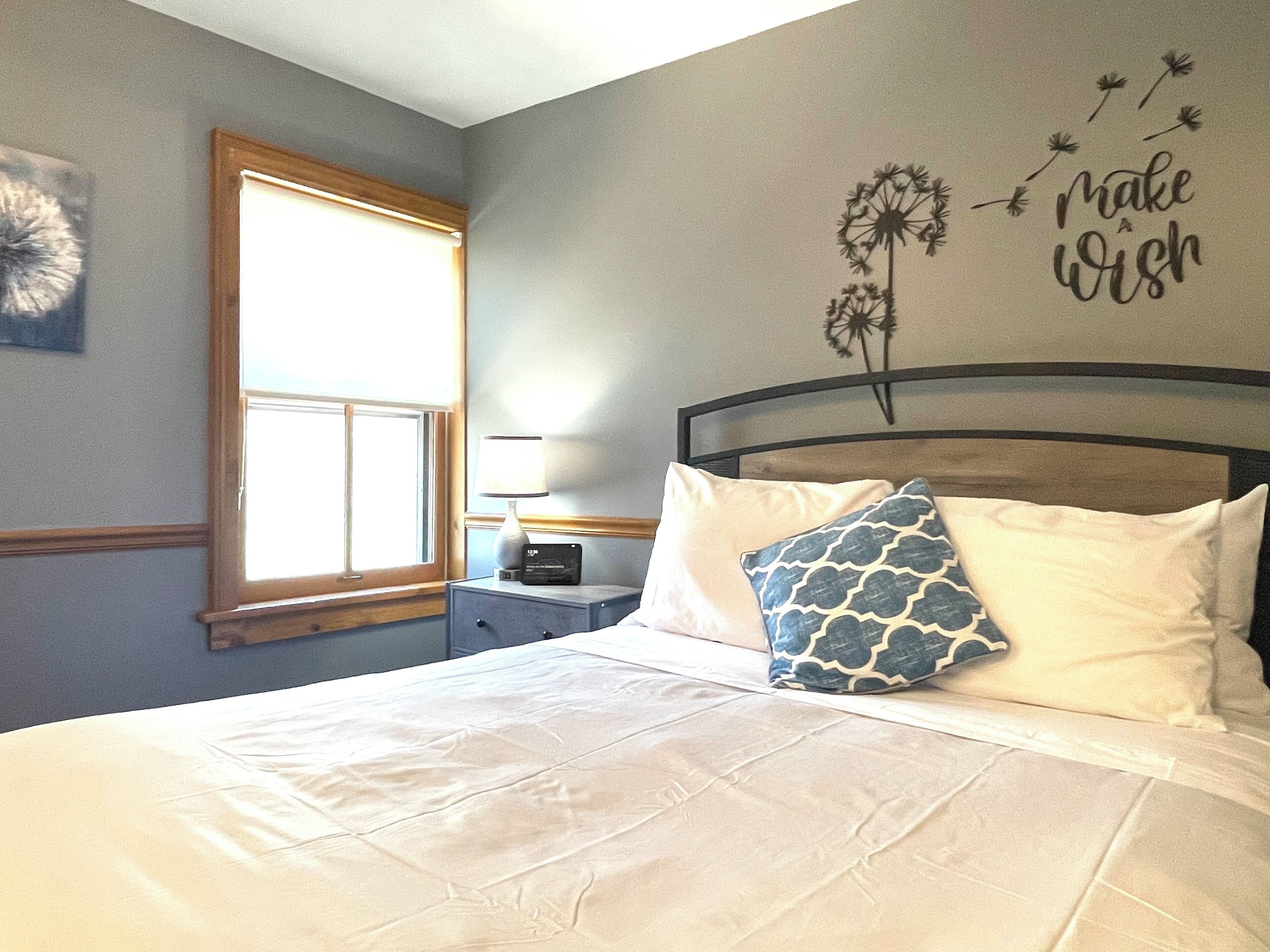 Une chambre à coucher avec un lit, des oreillers, une table de chevet, une lampe, une fenêtre et un élément de décoration murale.