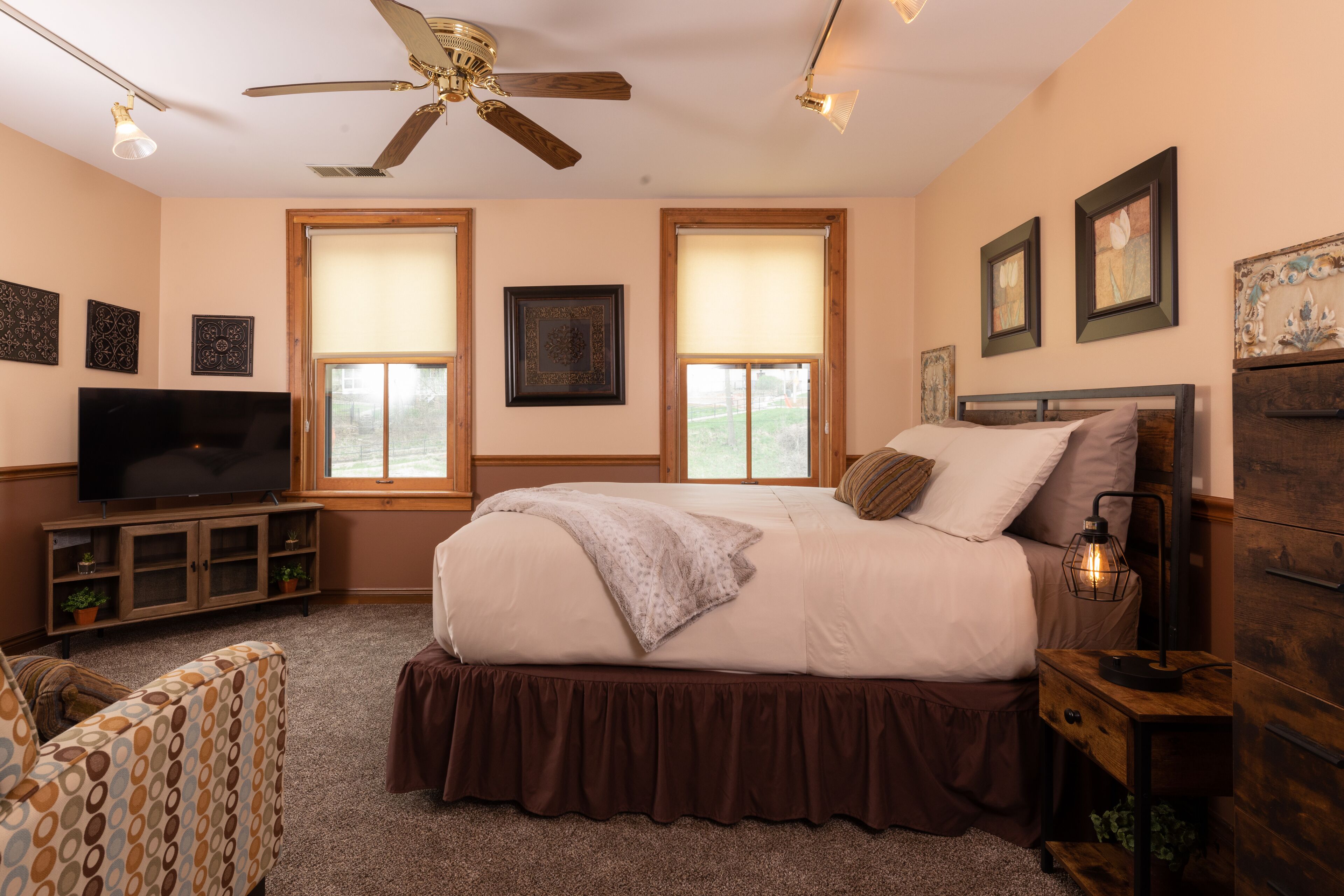 A bedroom with a large bed, a ceiling fan, two windows with blinds, a TV on a wooden cabinet, and a nightstand with a lamp.