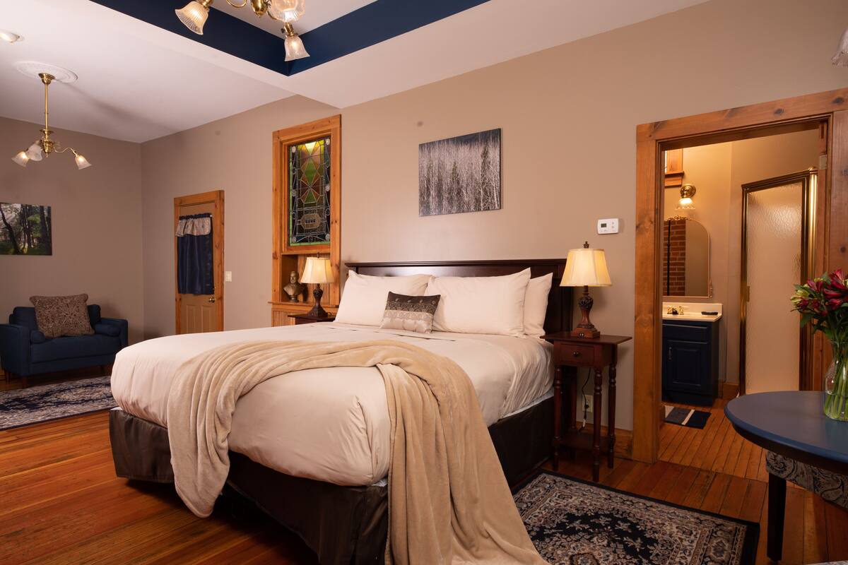 A bedroom with a large bed, a nightstand, a dresser, a blue chair, and a dining table with a vase of flowers.