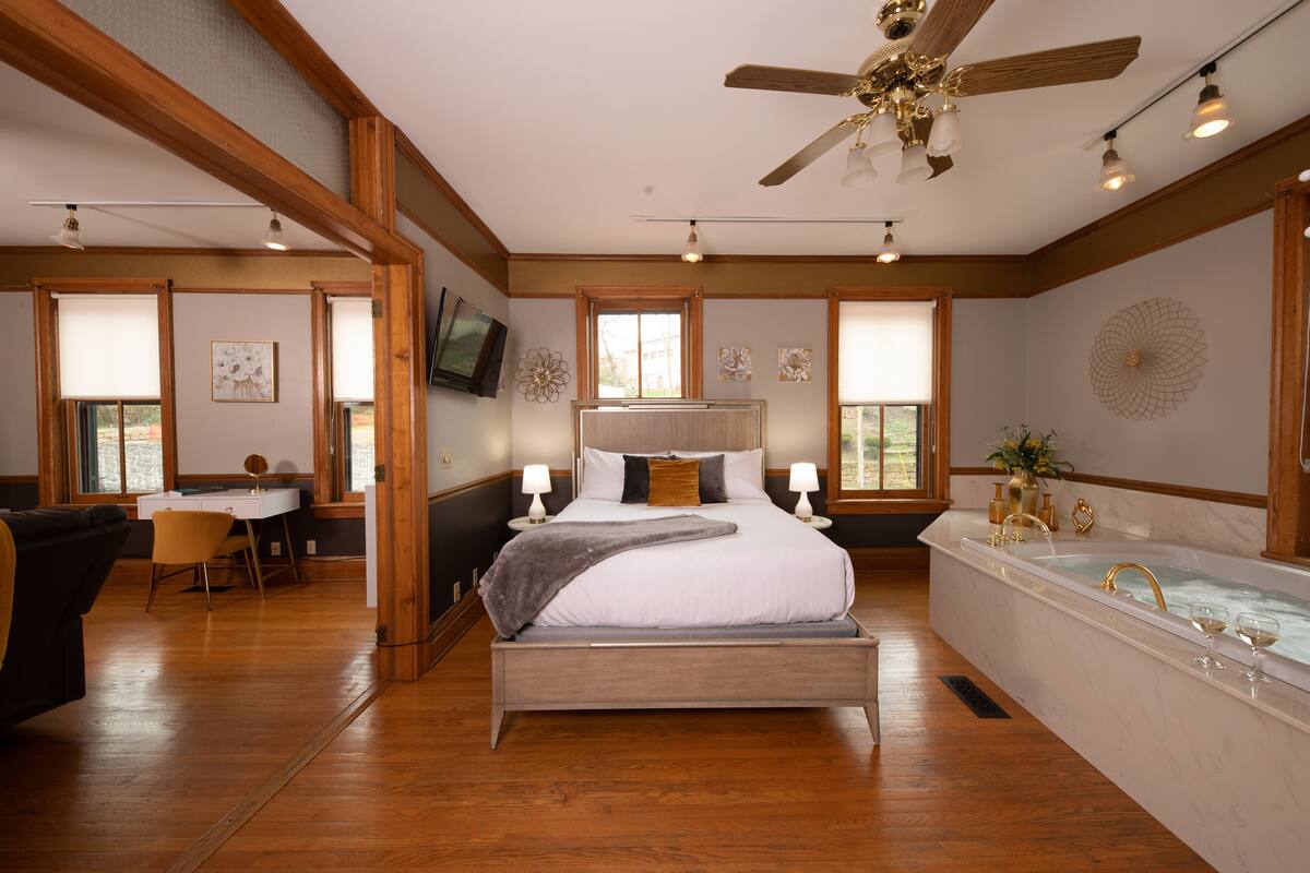 A spacious bedroom with a large bed, a sitting area with a desk, and a separate bathroom with a bathtub.