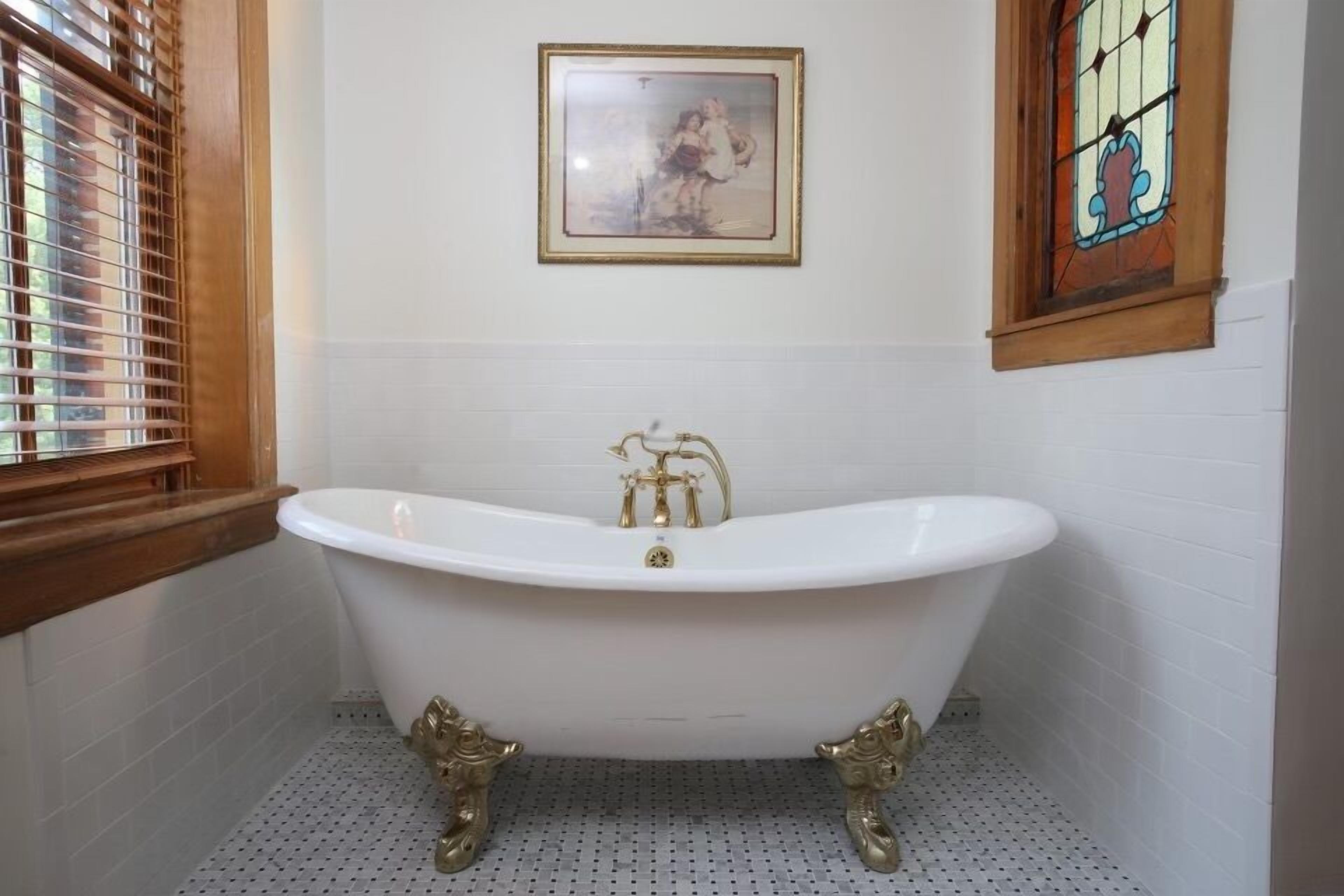 Une salle de bain avec une baignoire sur pieds, des stores en bois aux fenêtres, un tableau encadré au mur et un vitrail.