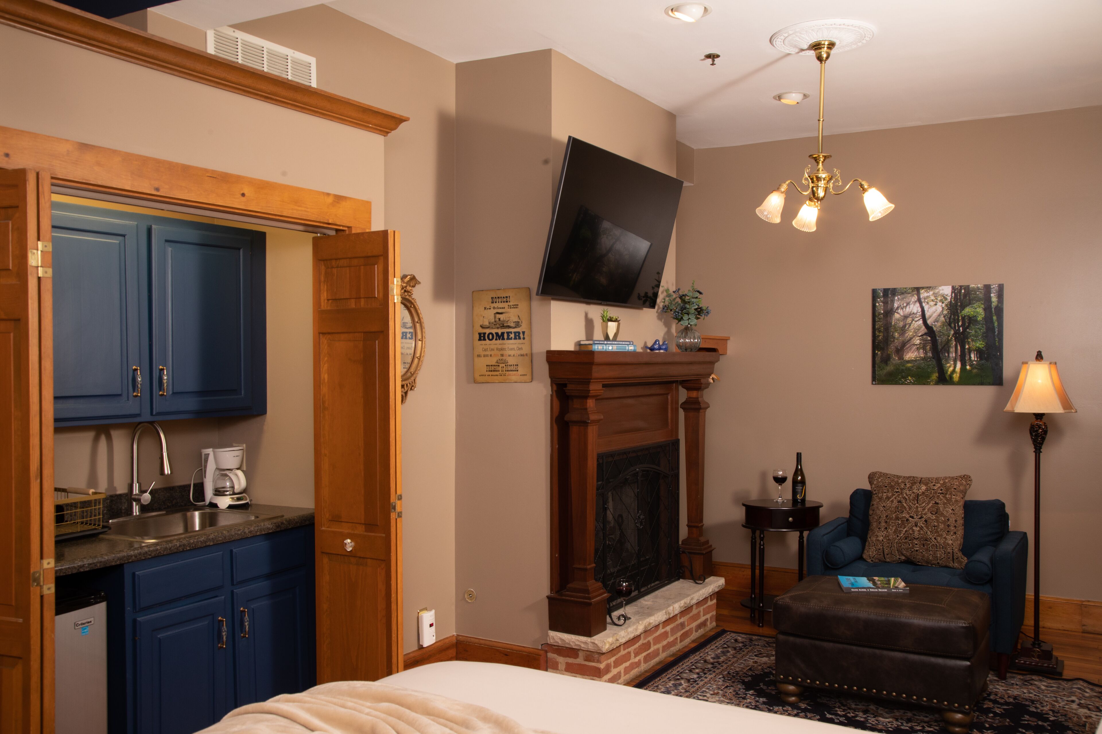 A hotel room with a kitchen area, a fireplace, a television mounted on the wall, a comfortable armchair, and a bedside table with a lamp.