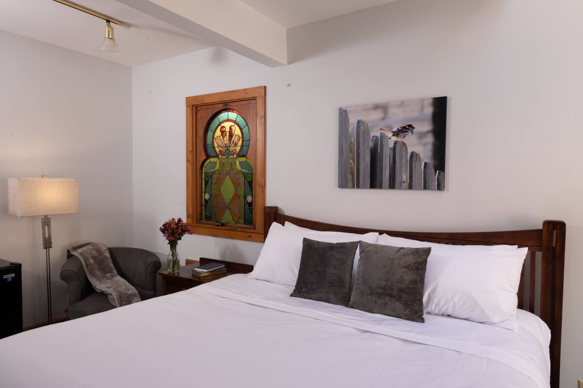 A neatly made bed with white linens and two gray pillows, a wooden headboard, a nightstand with a lamp and a book, a chair with a gray throw, a stained glass window, and a painting of birds on a fence.