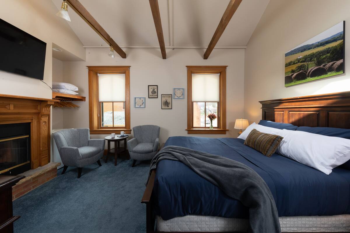 A hotel room with a large bed, a chair, a small table, a fireplace, and two windows.