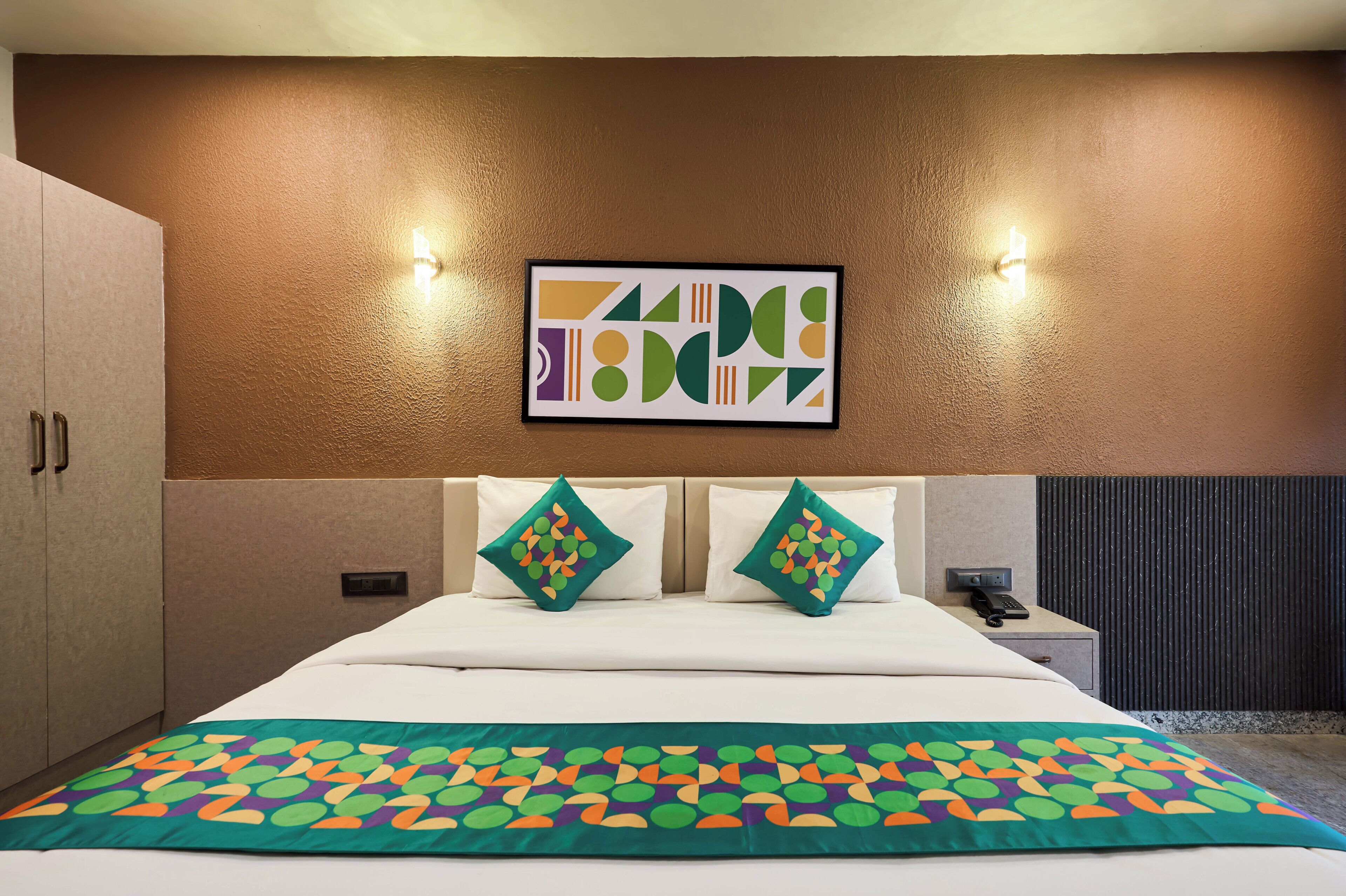 Ein Hotelzimmer mit Bett, zwei Kissen, einem Wandbild und einem Nachttisch mit Telefon, sowie einer Wand mit Strukturputz.