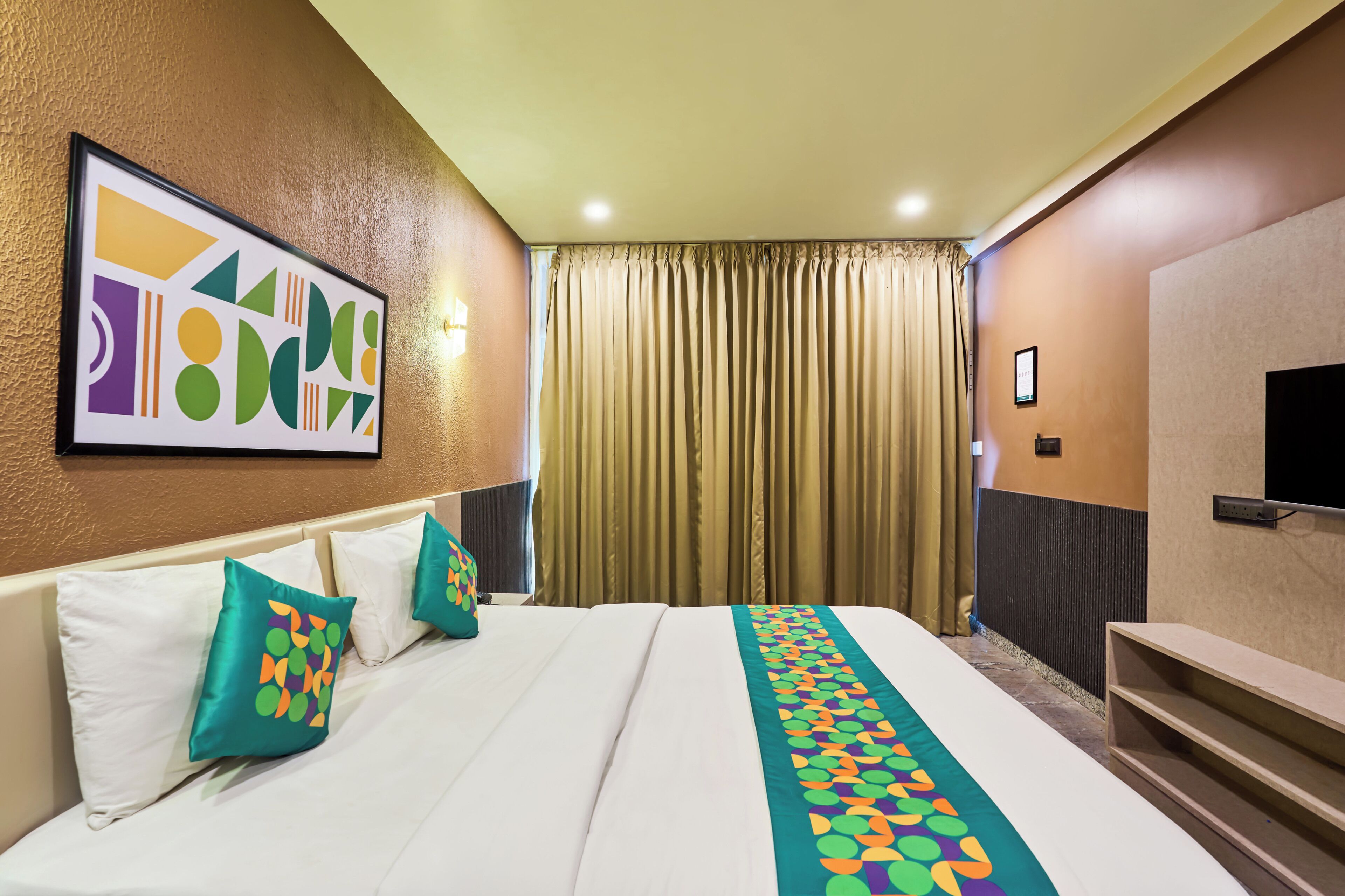 Una habitación de hotel con cama, almohadas, televisor, cortinas y una obra de arte en la pared.