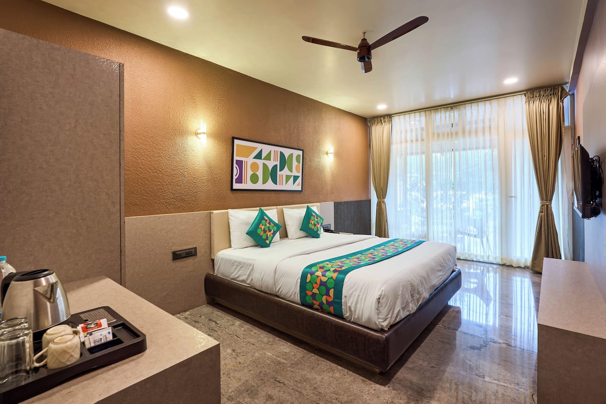 Habitación de hotel con cama, ventilador de techo, ventana con cortinas, televisor montado en la pared y bandeja con cafetera y vasos.