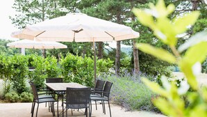 Une terrasse extérieure avec une table et des chaises, protégée par un grand parasol, et entourée de verdure et d’arbres.