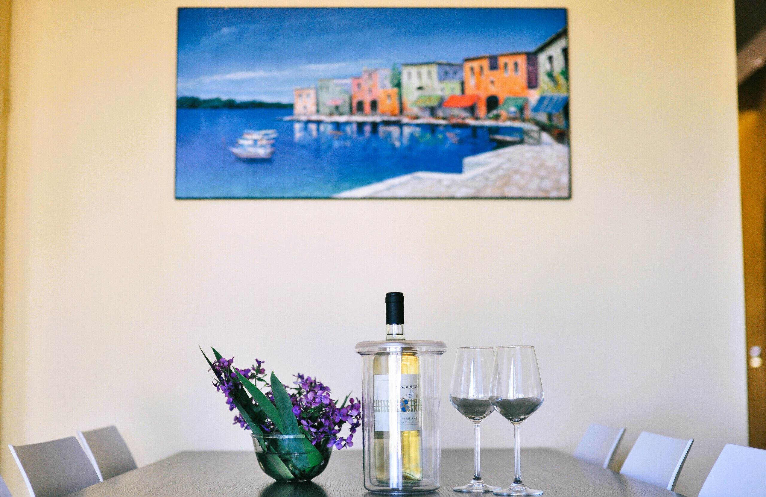 A dining table with a bottle of wine, two glasses, and a bowl of flowers.