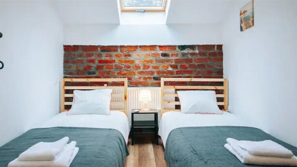 Ein Zimmer mit zwei Betten, einer Backsteinwand, einem Oberlicht und einem kleinen Tisch mit einer Lampe.