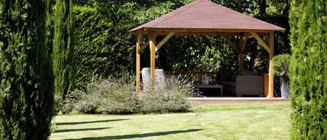 A gazebo with a wooden structure and a red roof, surrounded by a well-maintained lawn and various shrubs.