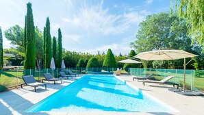 Une piscine entourée de chaises longues et de parasols, bordée de grands arbres et surplombée par un ciel dégagé.