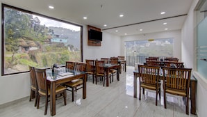 Un comedor con mesas y sillas de madera, ventanas amplias con vistas a una colina y un televisor montado.