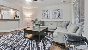 Ein modernes Wohnzimmer mit einem grauen Sofa, einem schwarzen Couchtisch und einem gemusterten Teppich. Es gibt einen Deckenventilator, eine Lampe und abstrakte Wandbilder.