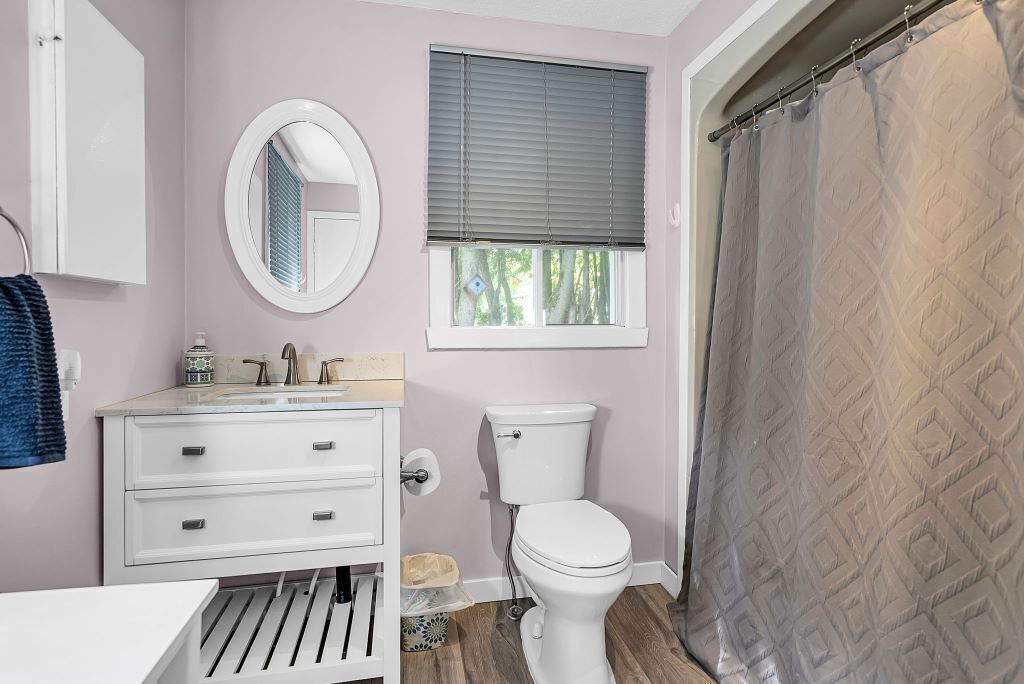 Une salle de bain équipée de toilettes, d’un lavabo, d’un miroir et d’une fenêtre avec des stores.